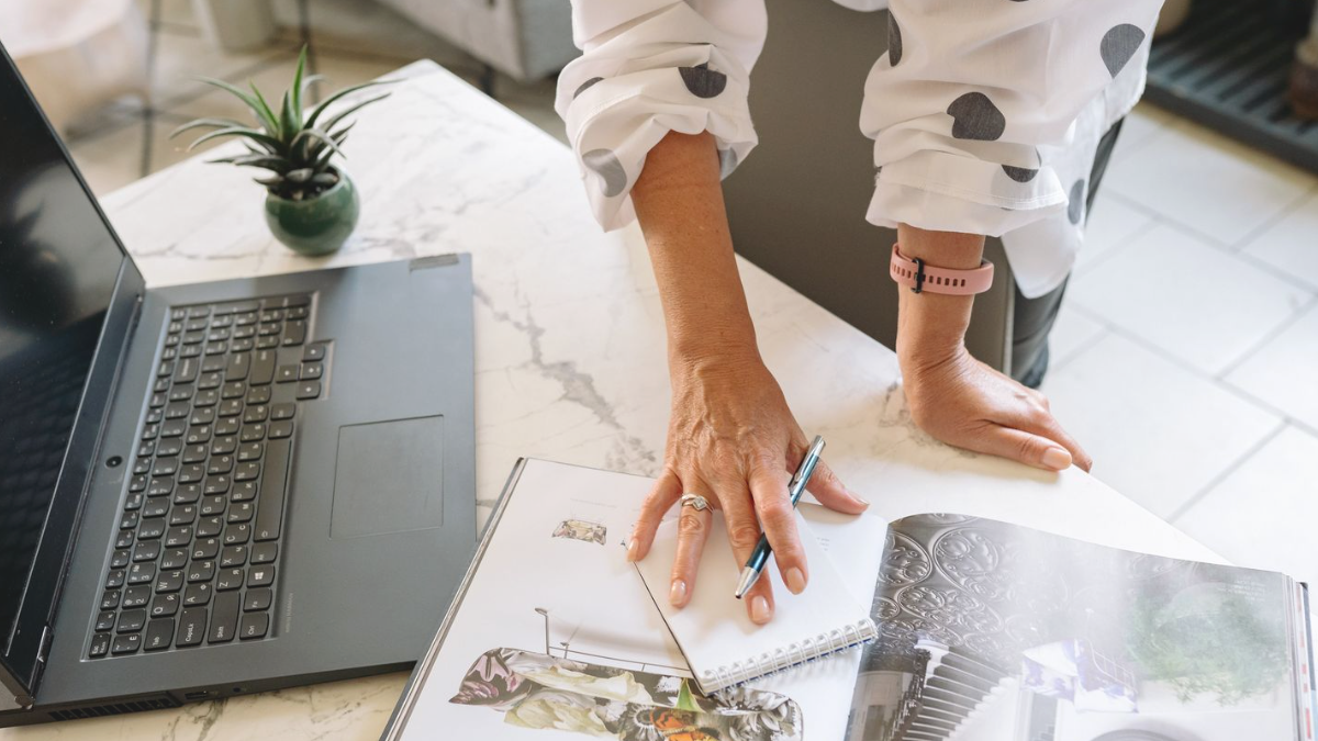 Mãos anotando em um caderno de planejamento ao lado do notebook, sobre uma mesa de mármore