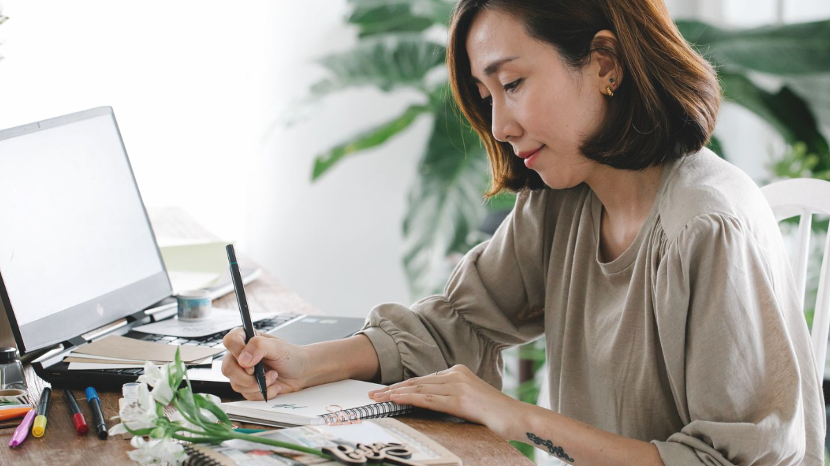 Mulher anotando em um caderno ao lado do notebook aberto, em mesa de madeira com plantas