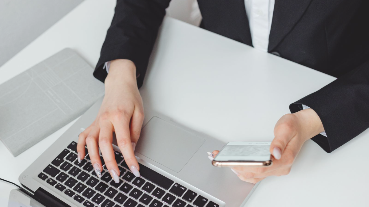 Mãos de uma profissional digitando no notebook enquanto segura o celular sobre a mesa