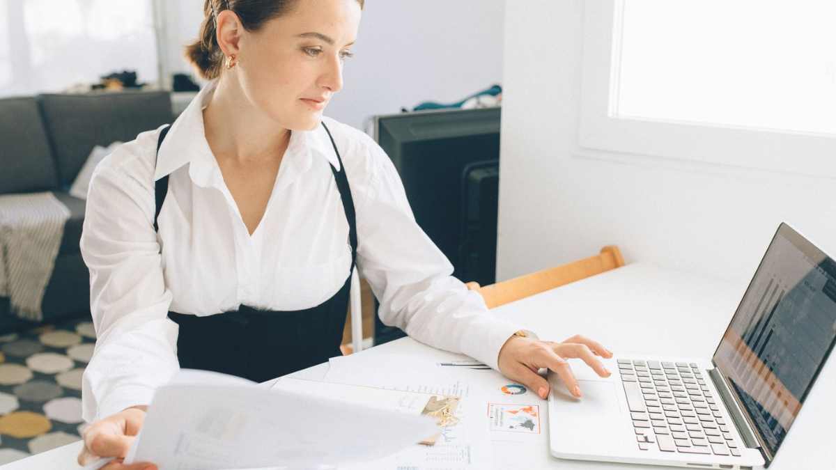 Mulher revisando relatórios impressos ao lado do notebook com gráficos na tela