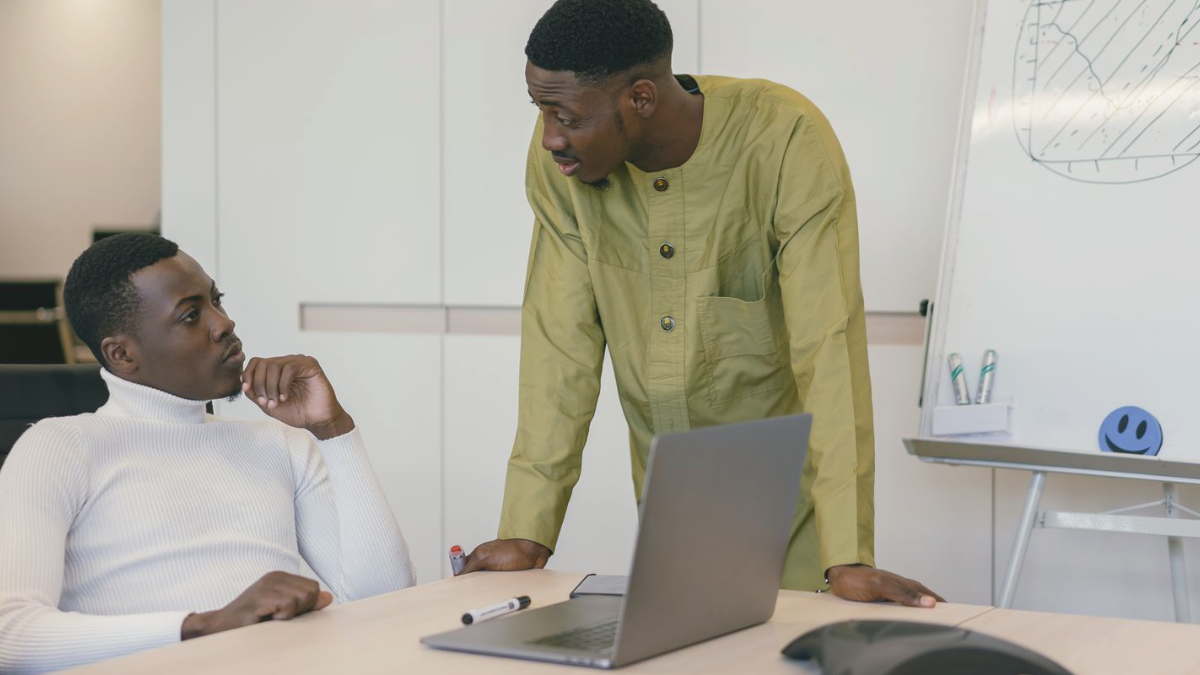 Dois colegas conversando em pé ao lado de um notebook em uma sala de reunião