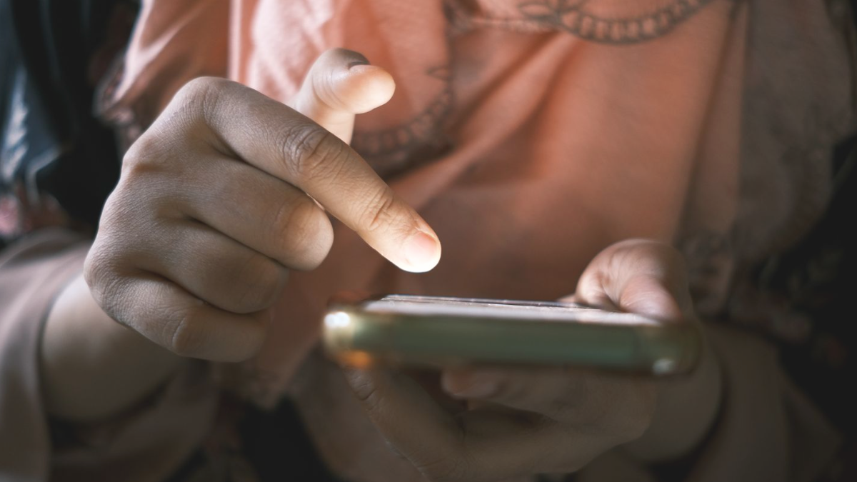 Close das mãos de uma mulher segurando um celular, com o dedo indicador esticado prestes a tocar a tela.