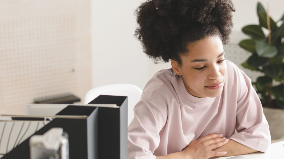 Jovem de blusa rosa sentada a uma mesa de escritório ao lado de um monitor, com um leve sorriso, com a tranquilidade que aparece quando você se posiciona e passa a ter critério para decidir.