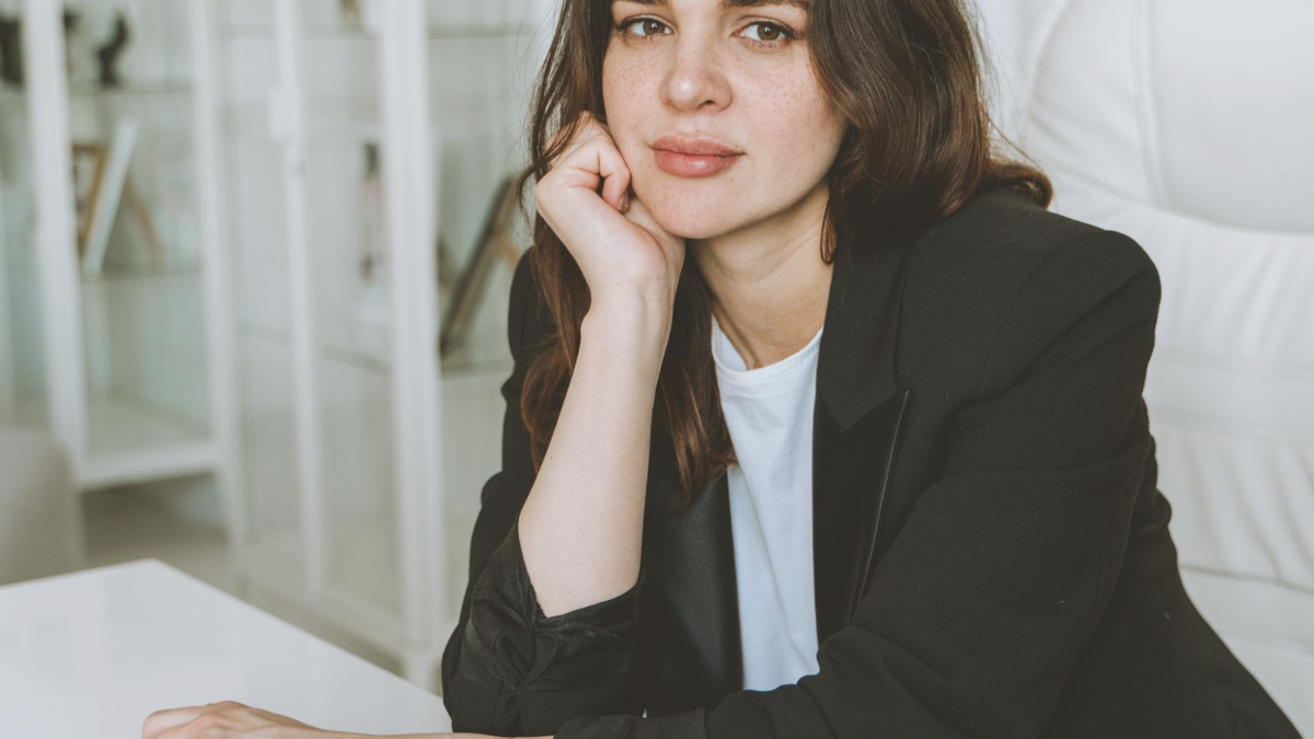 Profissional de blazer preto sentada a uma mesa clara, com o queixo apoiado na mão, pensando em como conseguir mais pacientes enquanto a colega menos qualificada tem a agenda lotada.